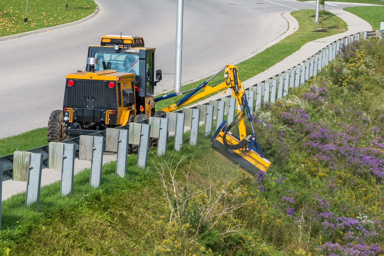 trackless vehicles boom flail mower attachment on sidewalk municipal tractor mowing grass in ditch rear view